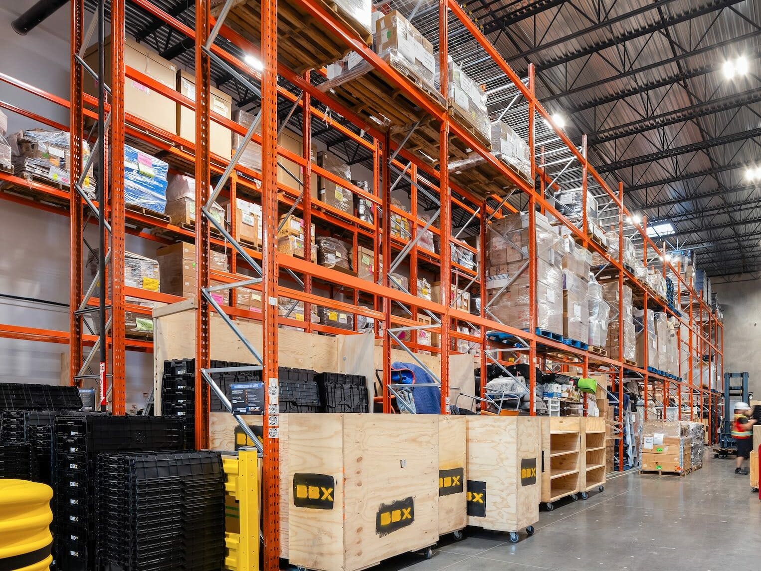 A warehouse. Pallets and crates are neatly organized, perfect for temporary storage needs. The ceiling's metal beams and bright lights illuminate the space efficiently.